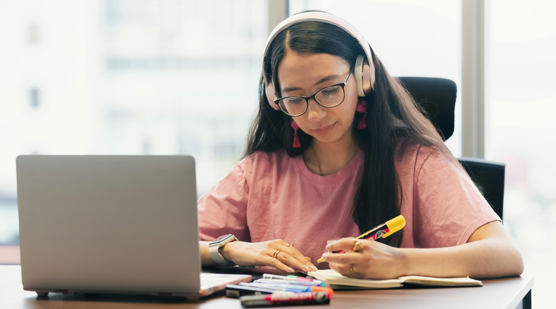 Young woman wearing headphones, writing in a notebook while studying at a laptop.
